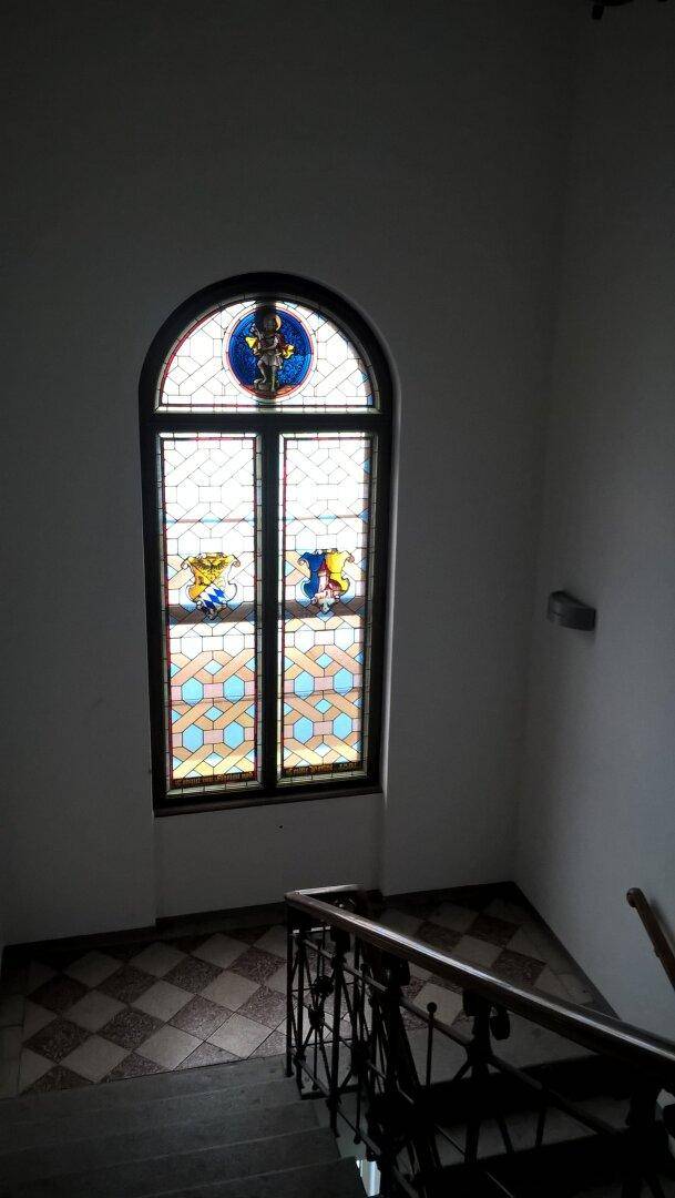Historisches Treppenhaus mit kunstvollem Buntglasfenster und schmiedeeisernem Geländer.