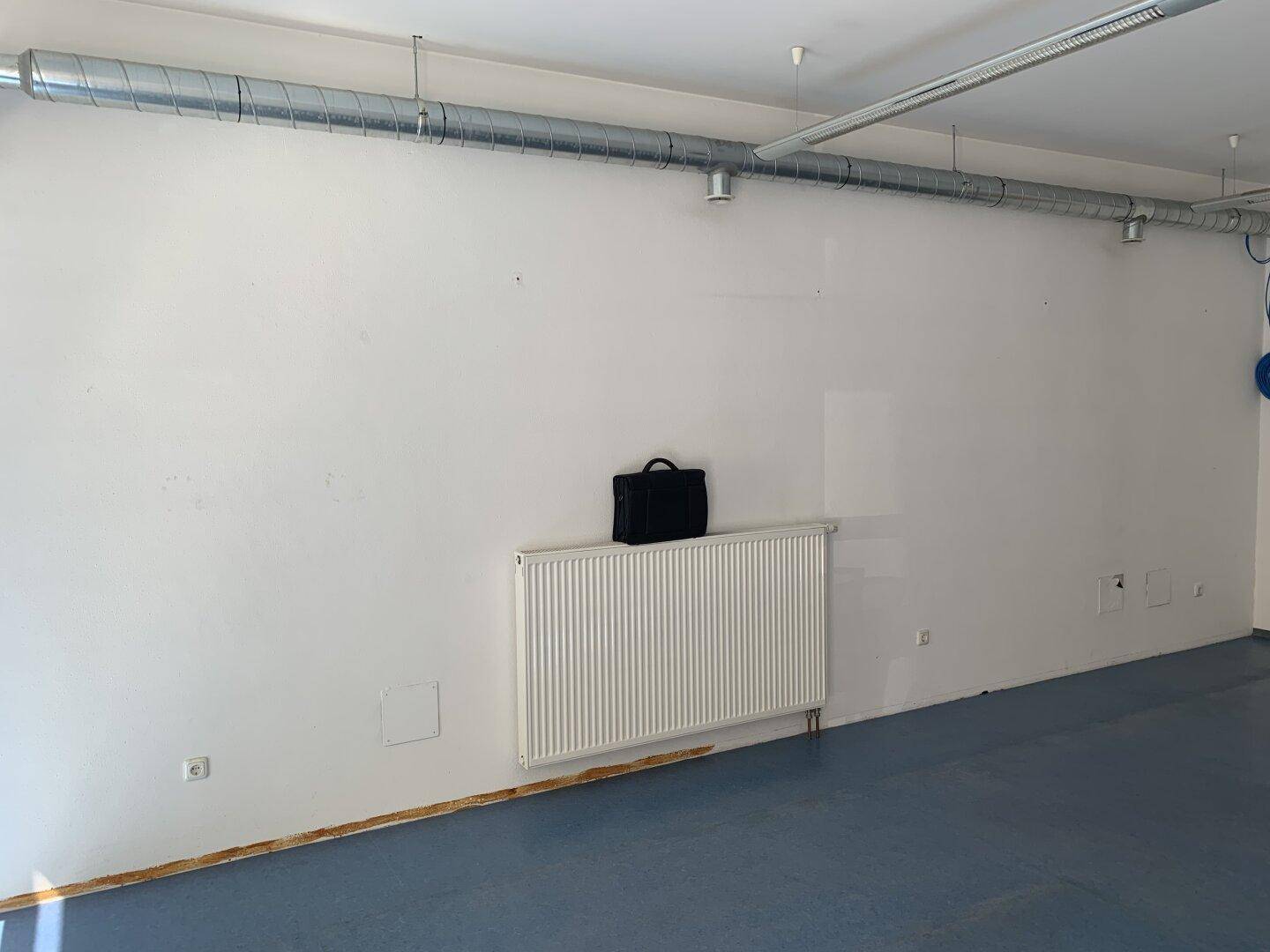 Leerer Gewerberaum mit weißen Wänden, Heizkörper und sichtbaren Lüftungsrohren an der Decke.