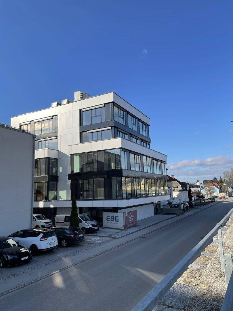 Modernes Bürogebäude an einer Straße mit Parkplätzen und klarer Architektur unter blauem Himmel.