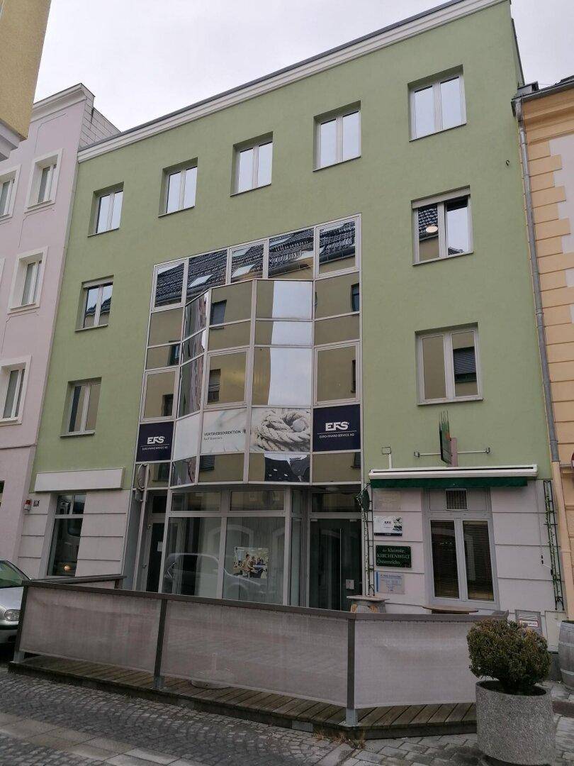 Gebäudefassade in Grün mit großen Glasflächen und einem kleinen Außenbereich mit Geländer.