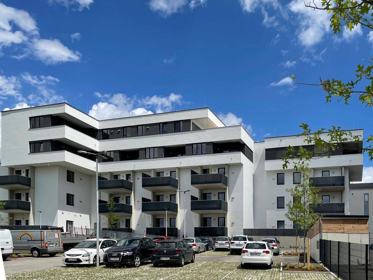 Modernes Mehrfamilienhaus mit mehreren Balkonen und Außenparkplätzen an einem sonnigen Tag.