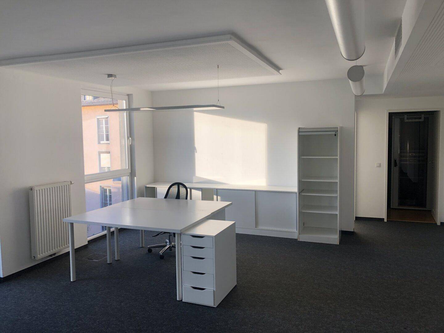Modernes Einzelbüro mit weißem Schreibtisch, Bürostuhl und Regalen, gut beleuchtet durch ein großes Fenster.