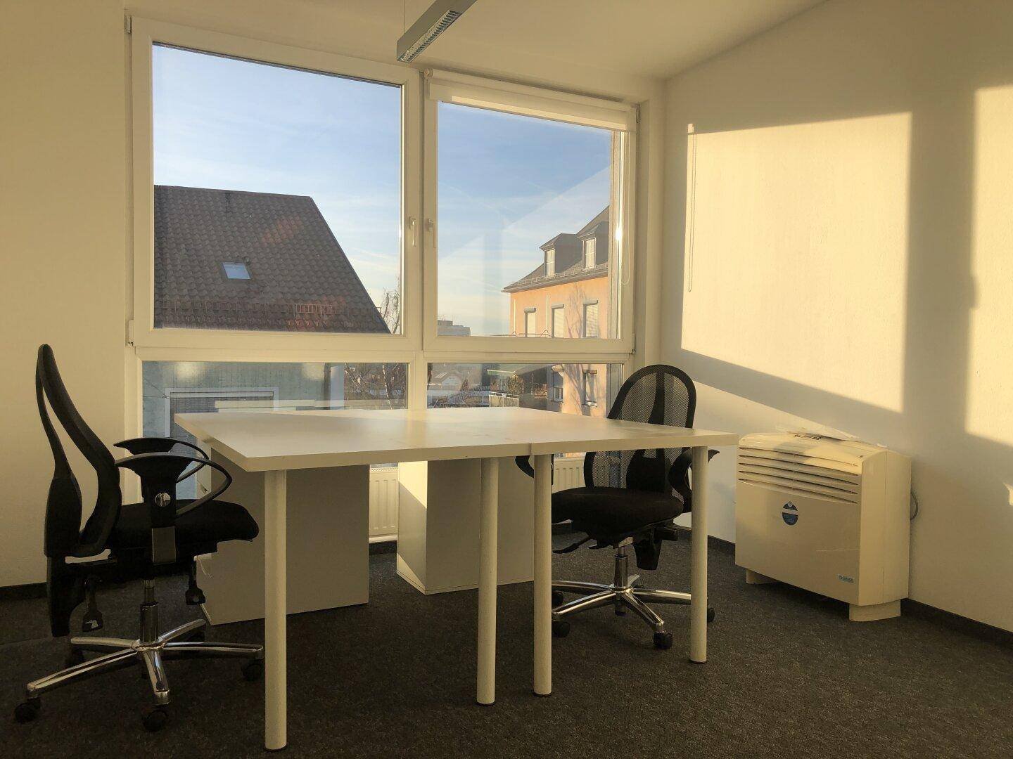 Helles Einzelbüro mit Schreibtisch und zwei Stühlen, ausgestattet mit einem Heizkörper und großen Fenstern.