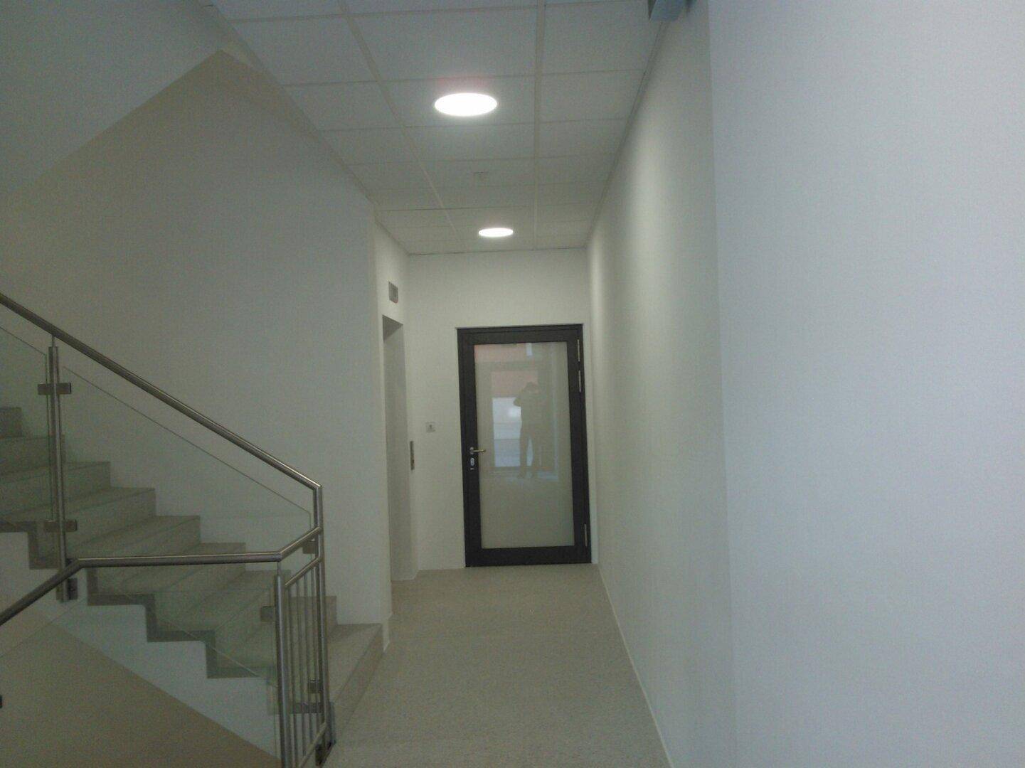 Heller Flur mit Treppe und Aufzug, ausgestattet mit modernen Einbauleuchten und Glasgeländer.