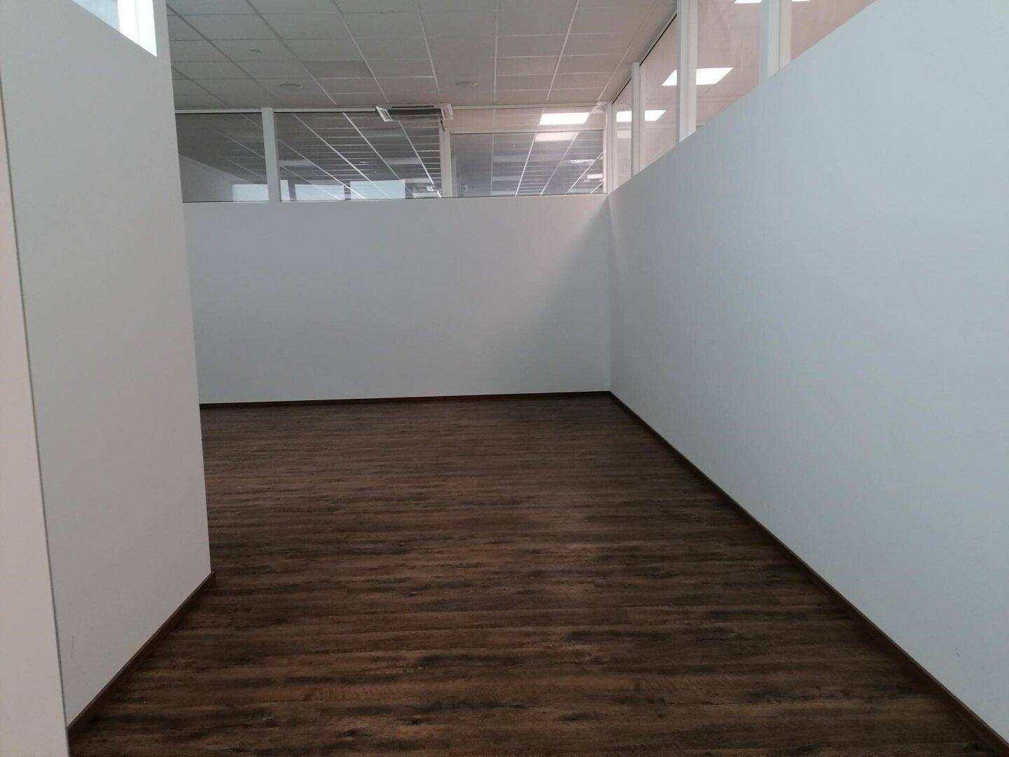 Heller Gewerberaum mit dunklem Holzboden, weißen Wänden und einer modernen Glasabtrennung im Hintergrund.