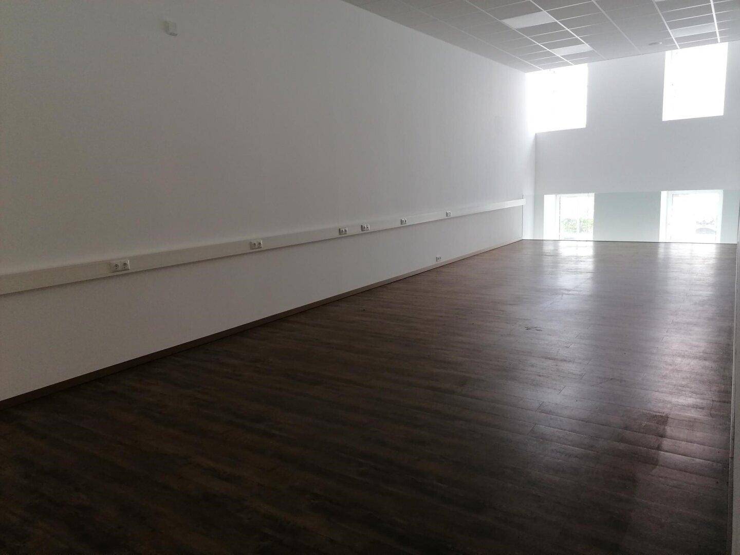 Geräumiger, leerer Gewerberaum mit dunklem Holzboden, weißen Wänden und hohen Decken mit integrierter Beleuchtung.
