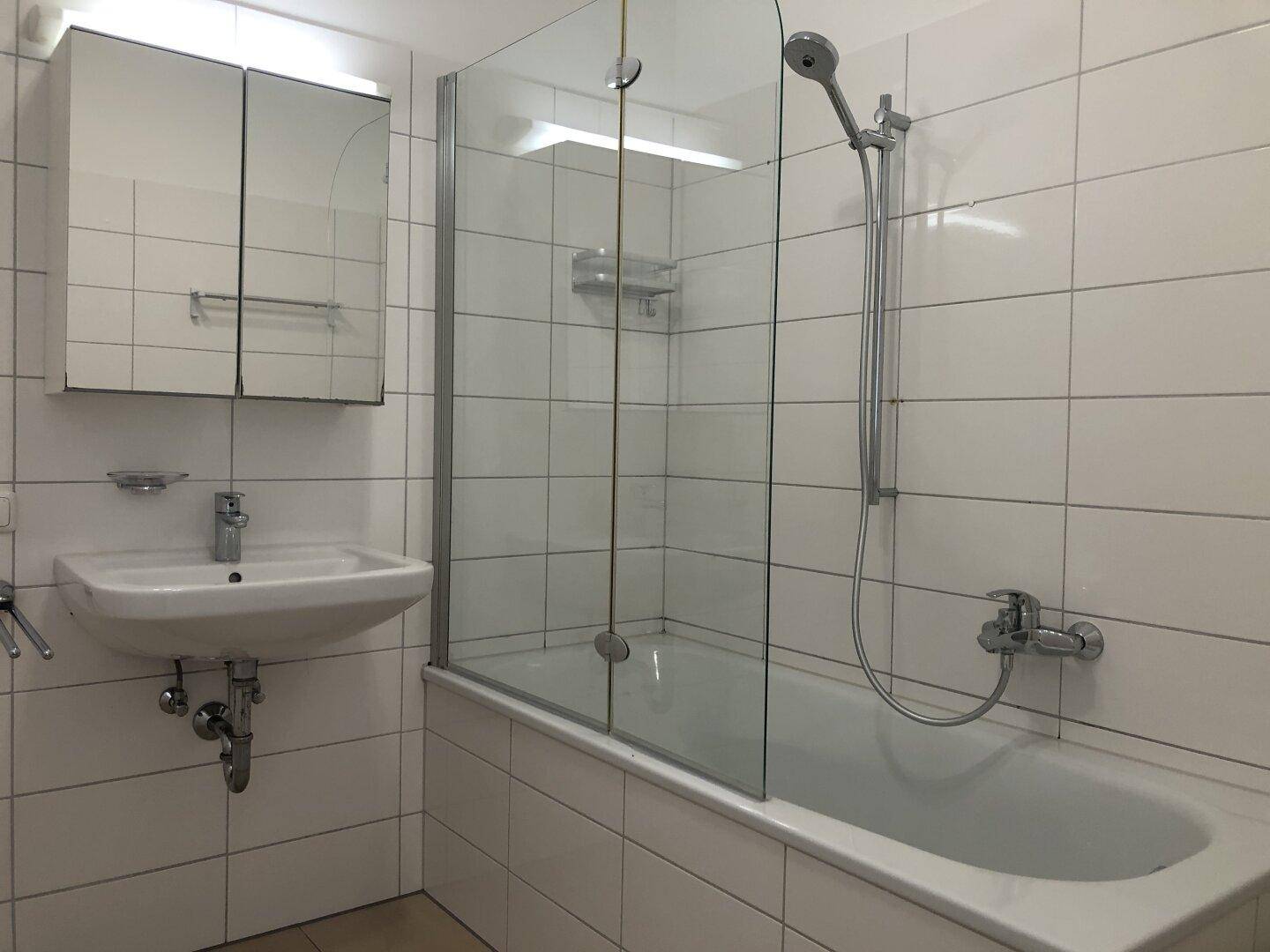 Helles Badezimmer mit weißen Fliesen, Badewanne mit Duschwand und Waschbecken.