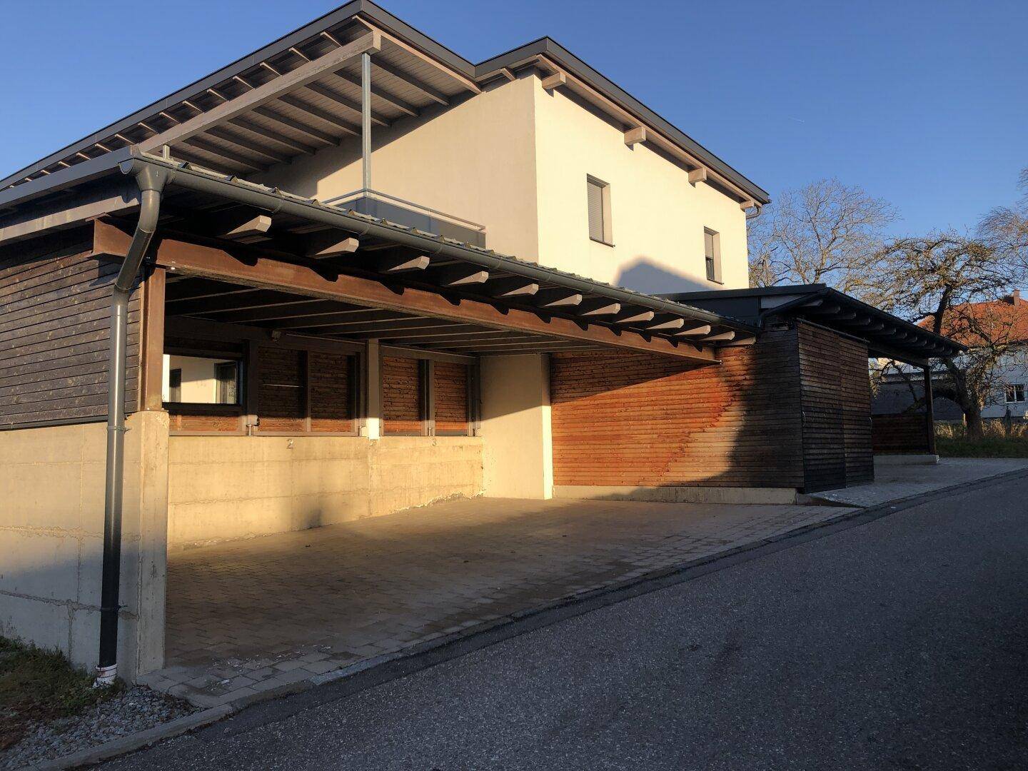 Carports mit Holzverkleidung und gepflasterter Zufahrt neben einem modernen Wohnhaus.