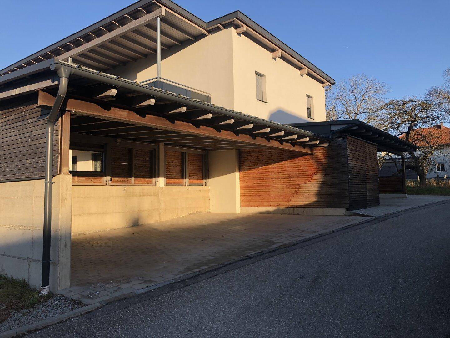 Moderne Carports mit Holzverkleidung und gepflastertem Bereich neben dem Wohngebäude.