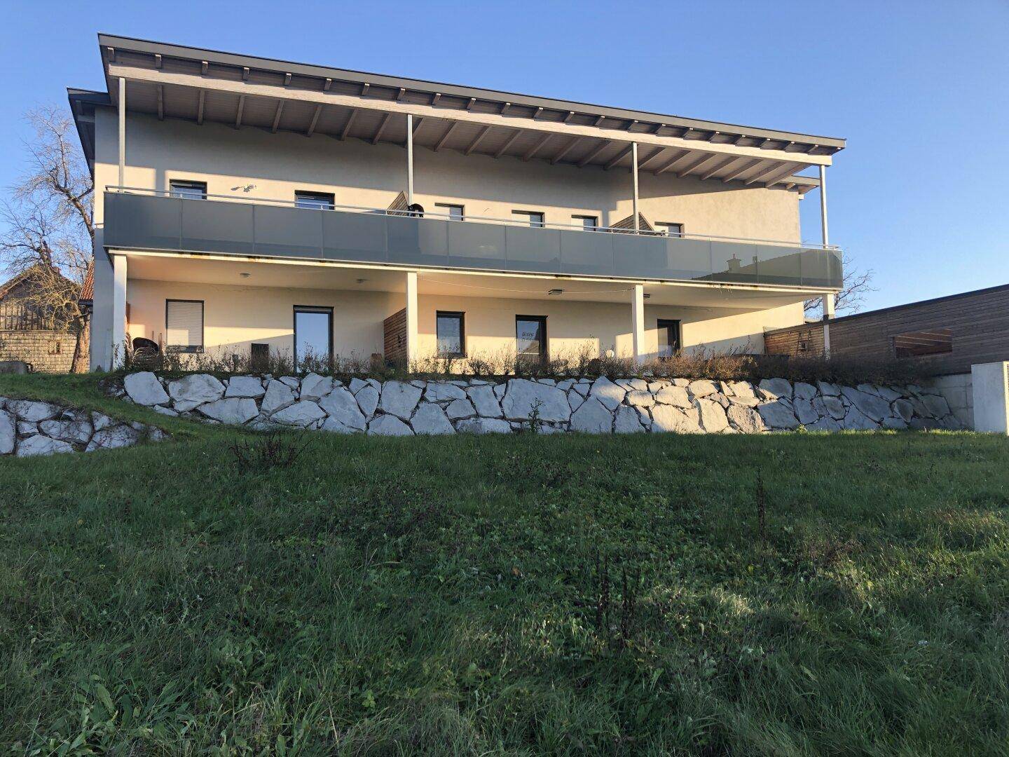 Ansicht eines modernen Mehrfamilienhauses mit Balkonen und einer Steinmauer vor einer grünen Wiese.