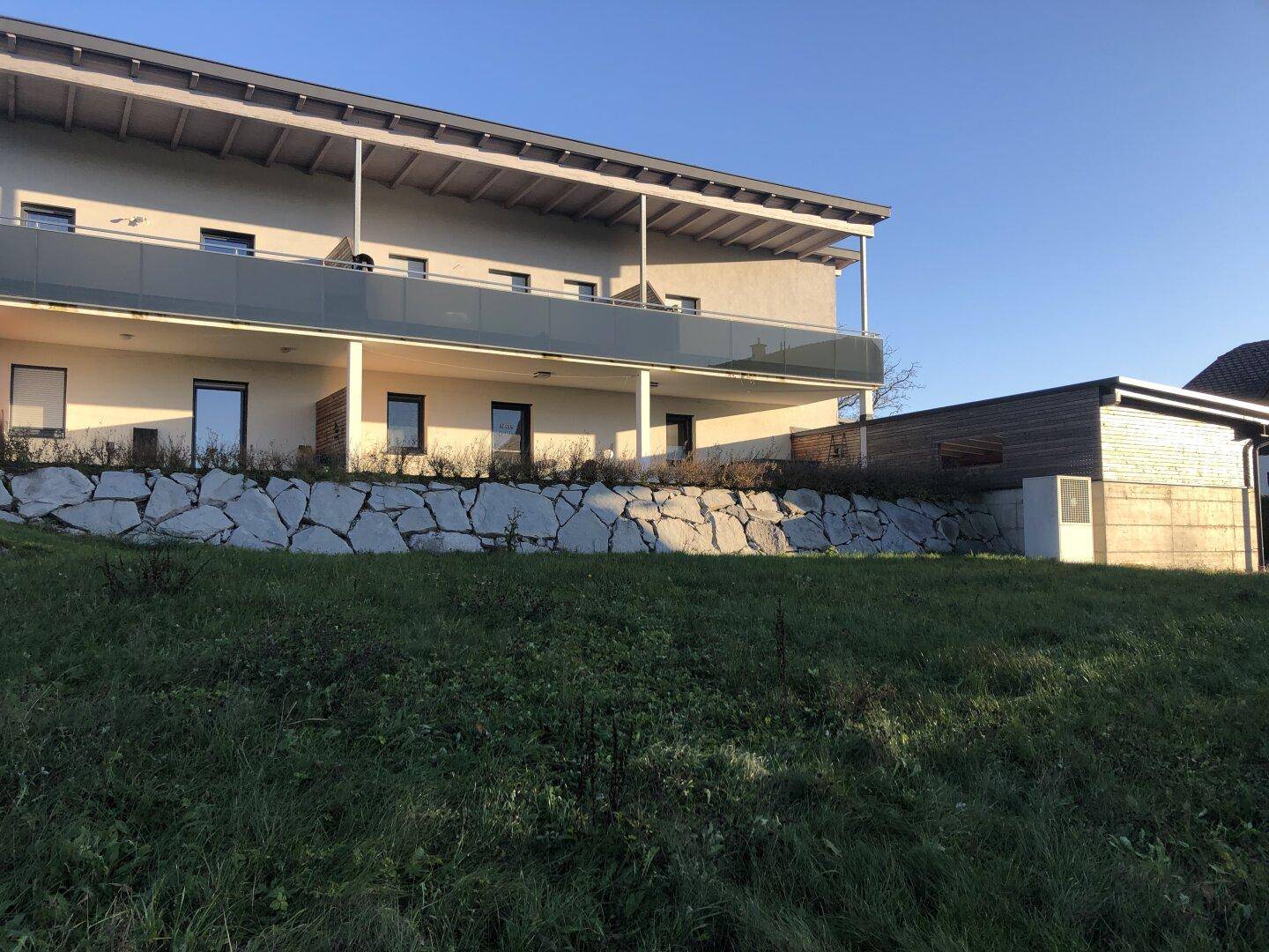 Modernes Gebäude mit Balkonen und einer Natursteinmauer, eingebettet in eine ländliche Umgebung.