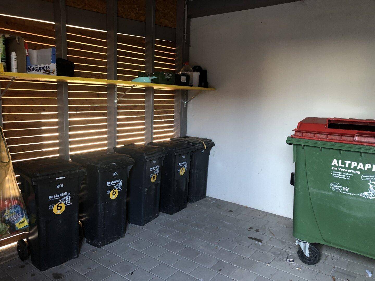 Abstellraum mit Mülltonnen und Regalen, ausgestattet mit Holzlamellen an der Wand.