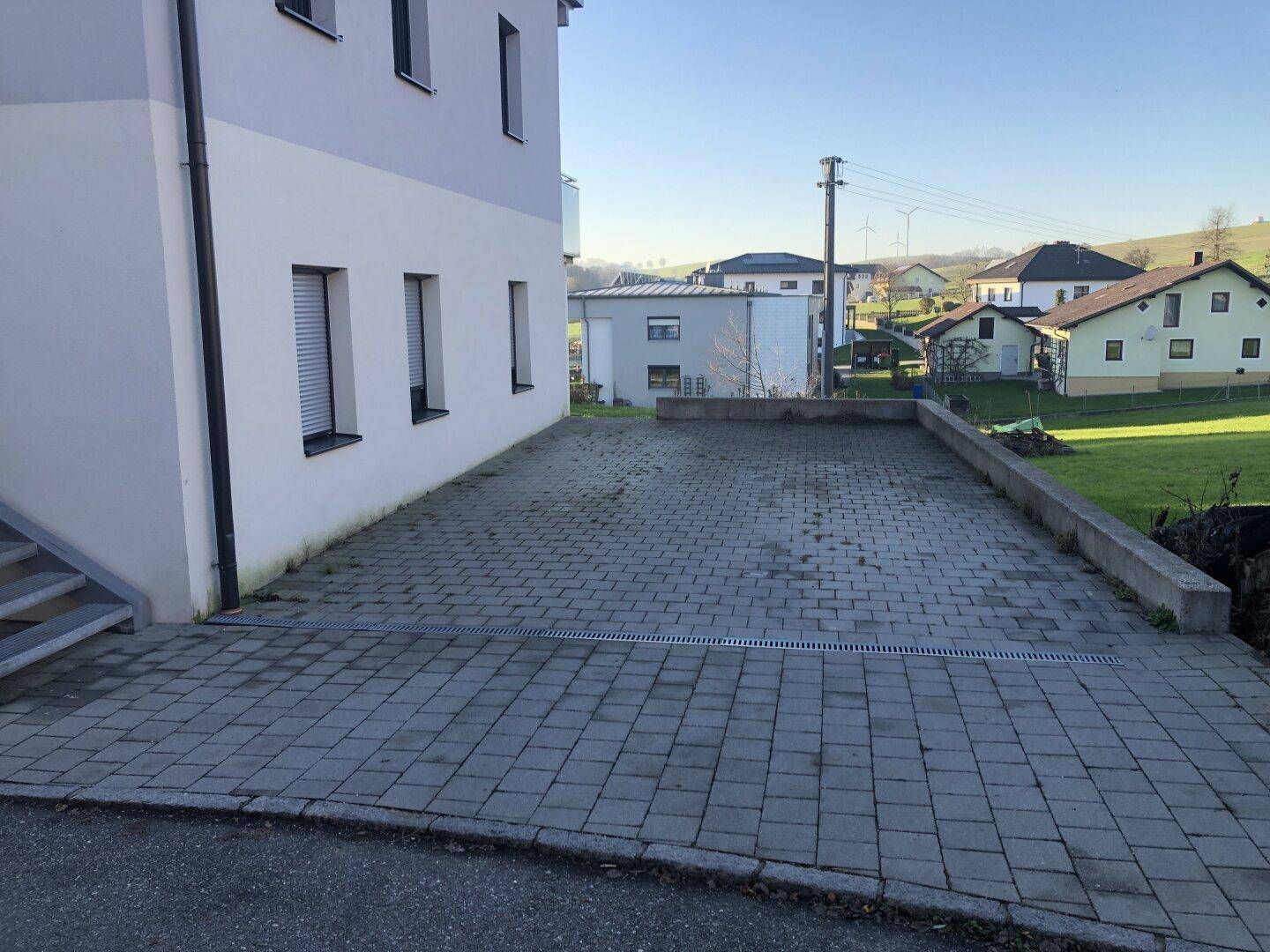 Großer gepflasterter Parkplatz neben dem Gebäude, umgeben von einer niedrigen Mauer.