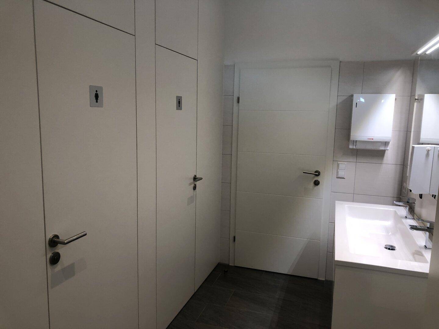 Öffentlicher Waschraum mit mehreren Toilettenkabinen, einem modernen Waschbecken und großen Spiegeln.