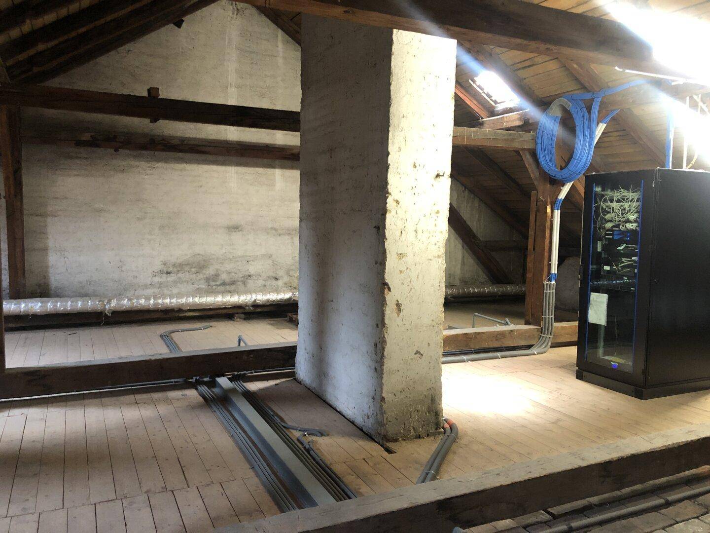 Dachboden mit sichtbaren Holzbalken, Holzboden und einem Serverschrank, der auf eine technische Nutzung hinweist.
