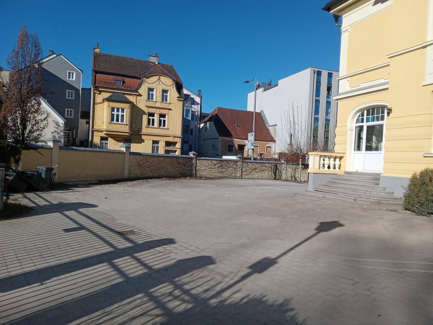 Außenbereich mit Schotterparkplatz und Blick auf umliegende Gebäude unter klarem, blauem Himmel.
