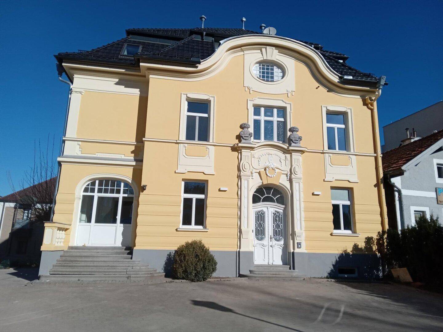 Historisches Gebäude mit gelber Fassade, weißen Fenstern und einem markanten Eingangsbereich unter blauem Himmel.