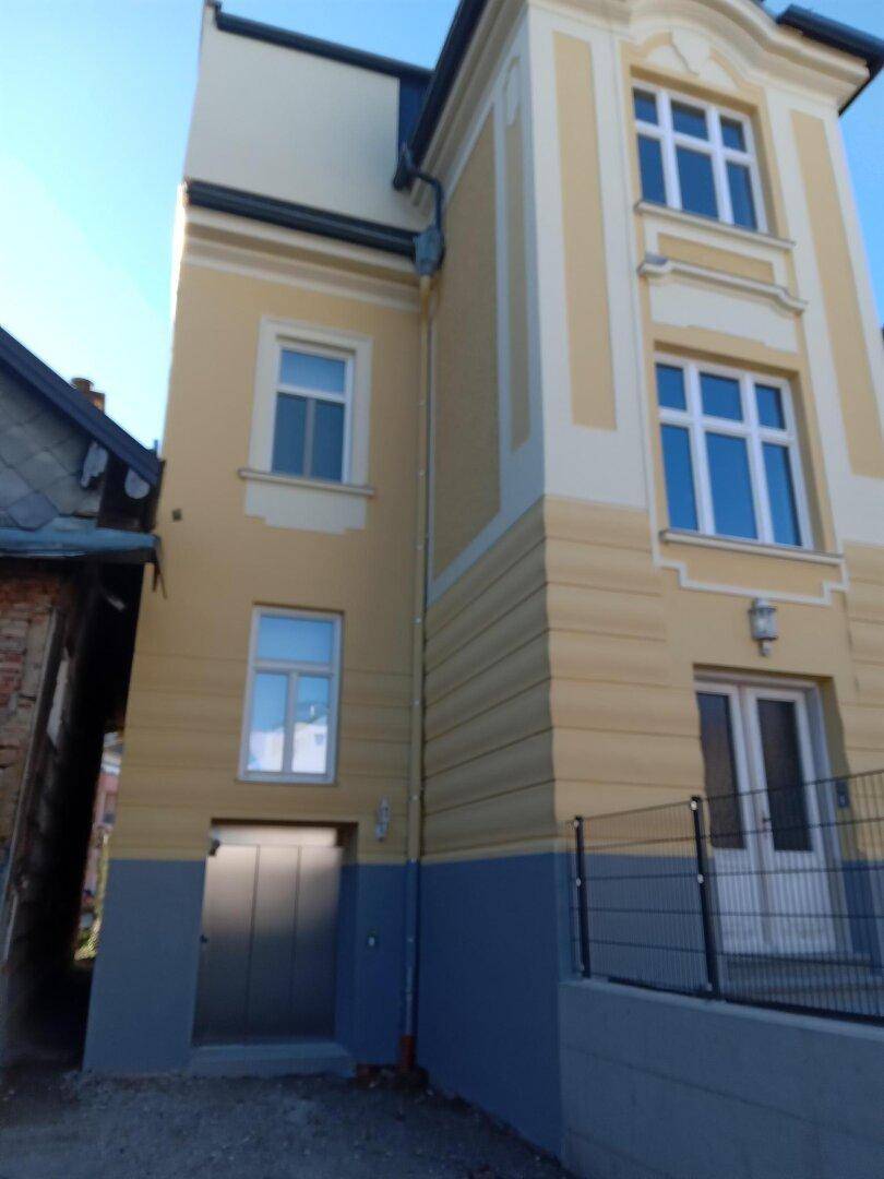 Seitenansicht des Gebäudes mit gelber und grauer Fassade, Fenstern und einem Eingang im Untergeschoss.