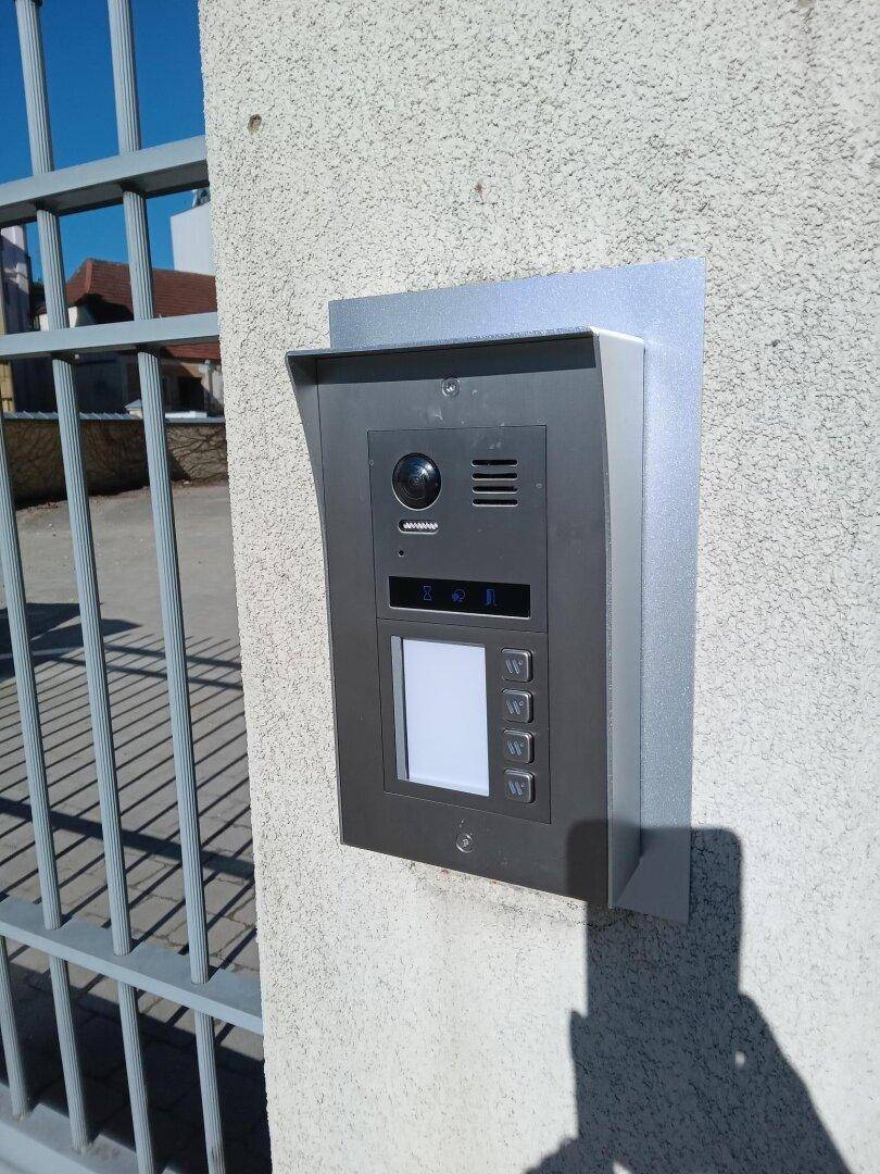 Moderne Gegensprechanlage mit Kamera und Tasten an einer Außenwand neben einem Metalltor.