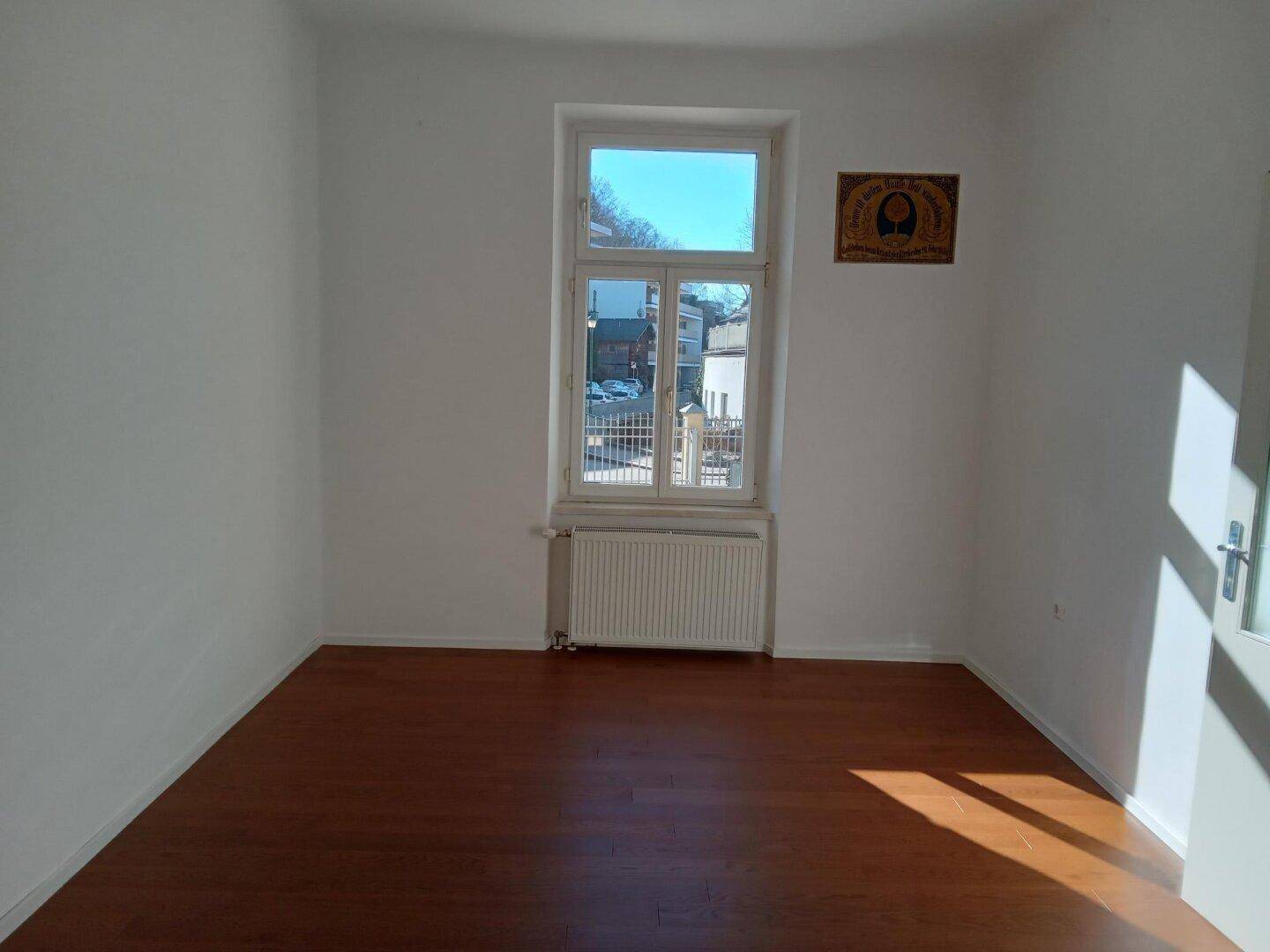 Leerer Raum mit Holzboden, weißen Wänden und einem großen Fenster, das viel Tageslicht hereinlässt.