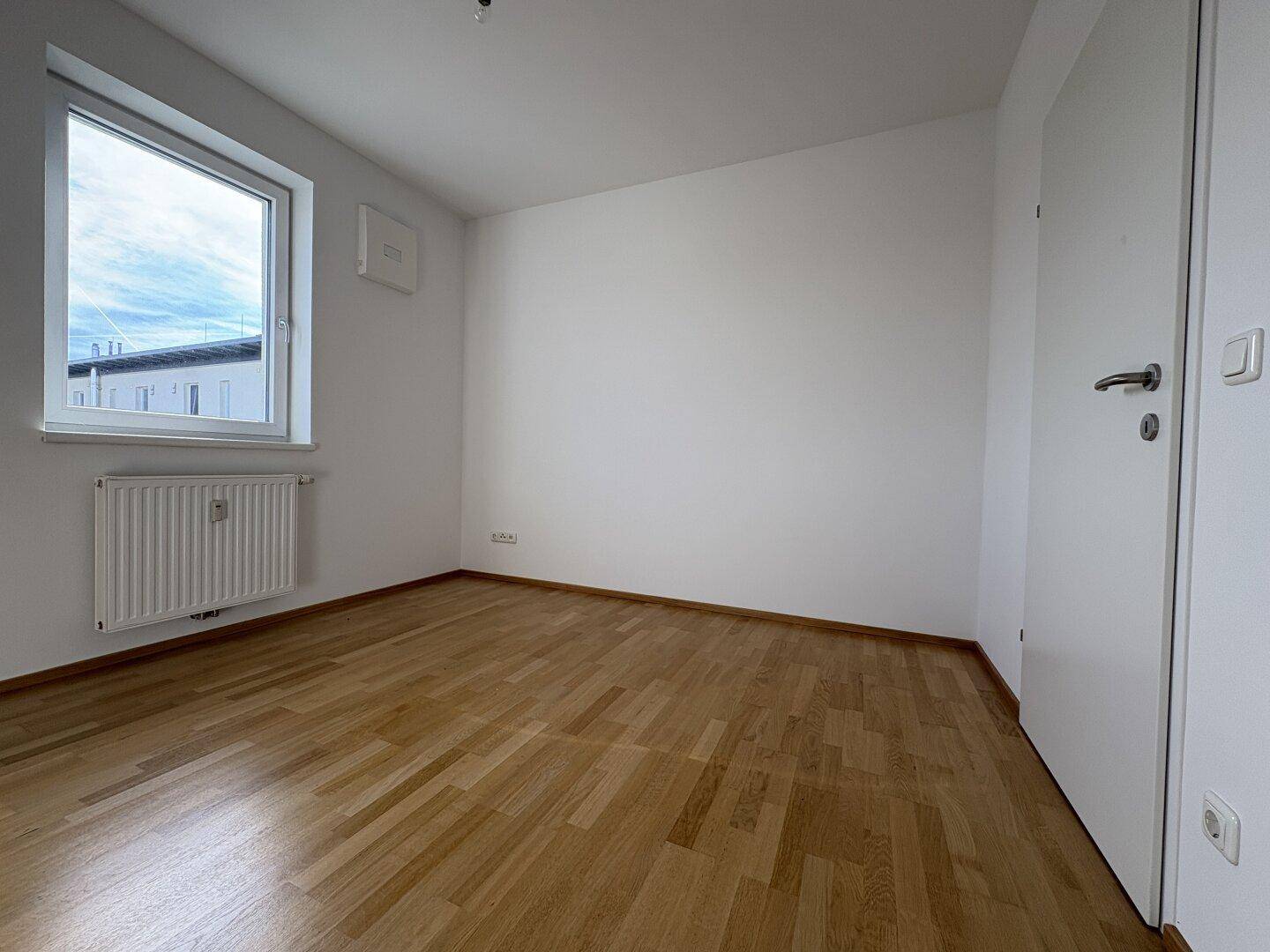Helles Zimmer mit Parkettboden und einem Fenster, das viel Tageslicht hereinlässt.