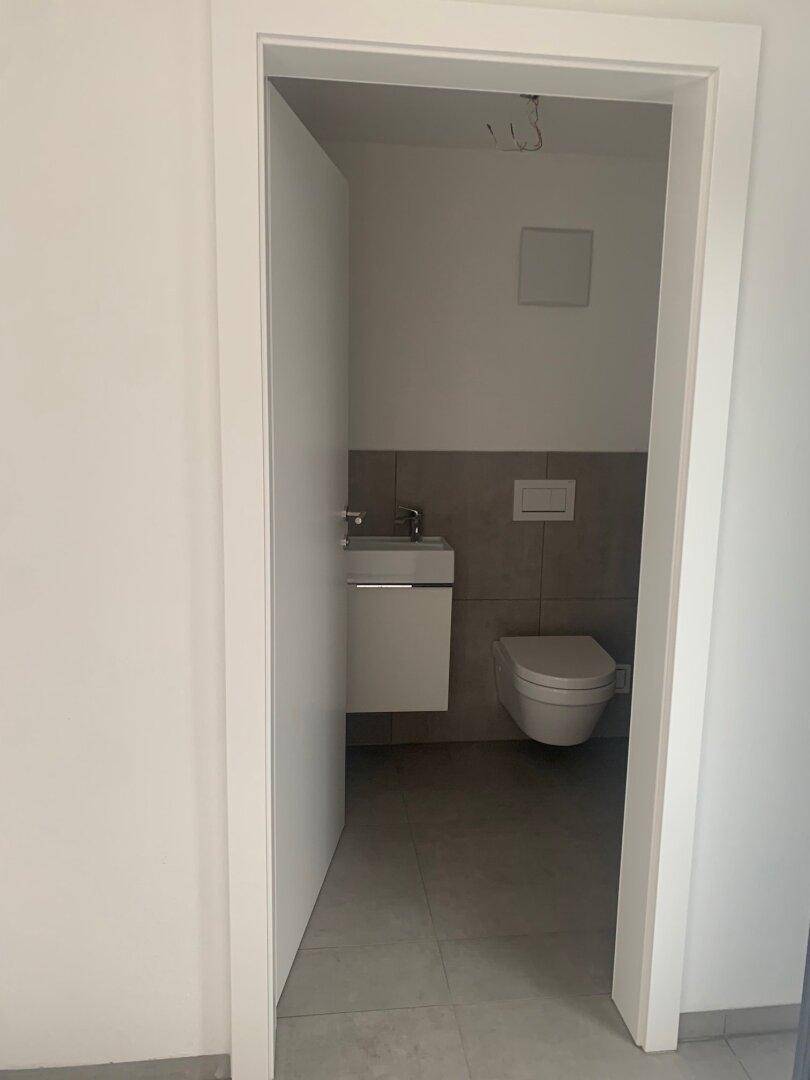 Modernes Gäste-WC mit wandhängender Toilette, kleinem Waschbecken und grauen Fliesen an Boden und Wänden.