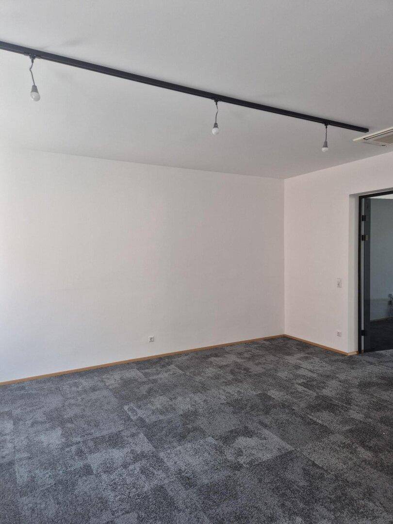 Geräumiger, leerer Büroraum mit grauen Teppichfliesen, weißen Wänden und moderner Deckenbeleuchtung.