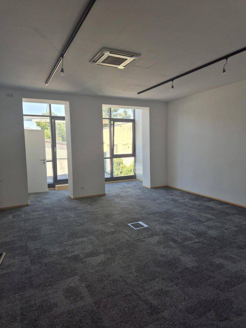 Geräumiger, leerer Büroraum mit grauen Teppichfliesen, großen Fenstern und Deckenbeleuchtung.