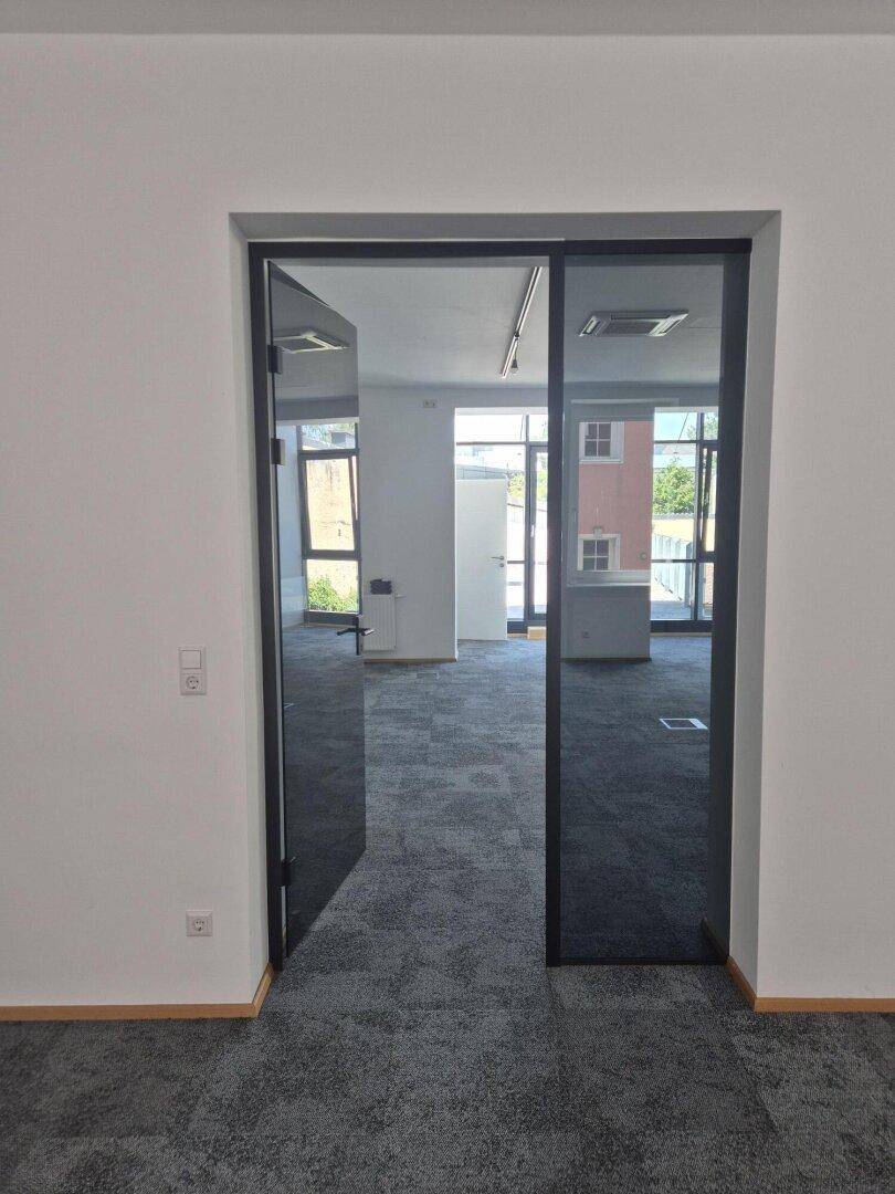 Blick durch eine moderne Glastür in einen hellen Büroraum mit grauen Teppichfliesen und großen Fenstern.