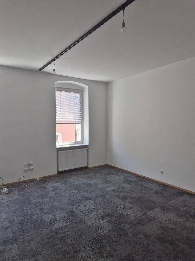 Leerer Büroraum mit grauen Teppichfliesen, weißen Wänden und einem Fenster mit Jalousien.