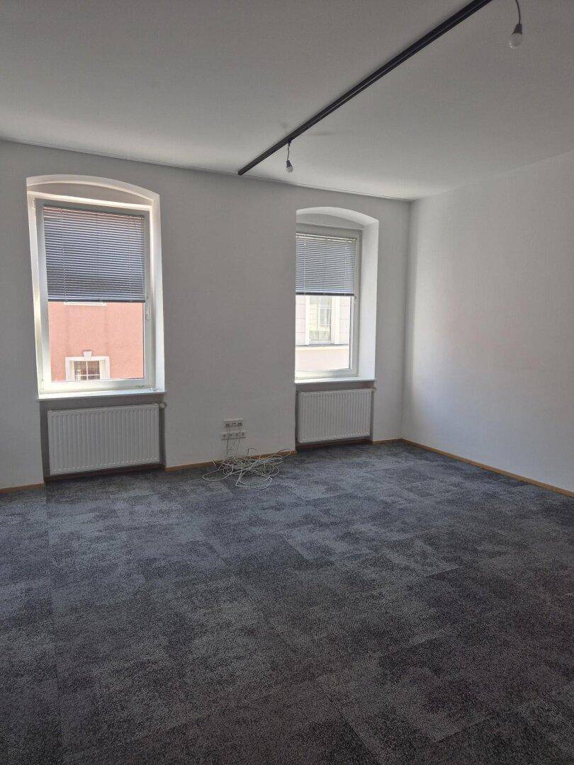 Heller Büroraum mit grauen Teppichfliesen, weißen Wänden und zwei Fenstern mit Jalousien.