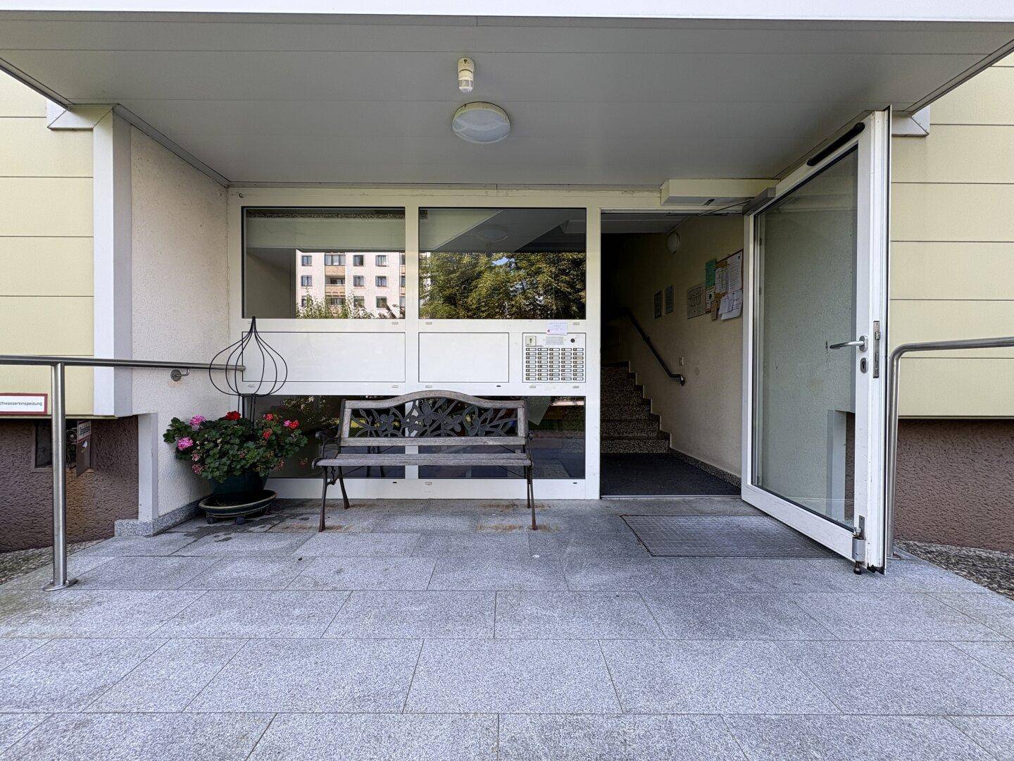 Eingangsbereich eines Mehrfamilienhauses mit Rampe, Sitzbank und offener Glastür.