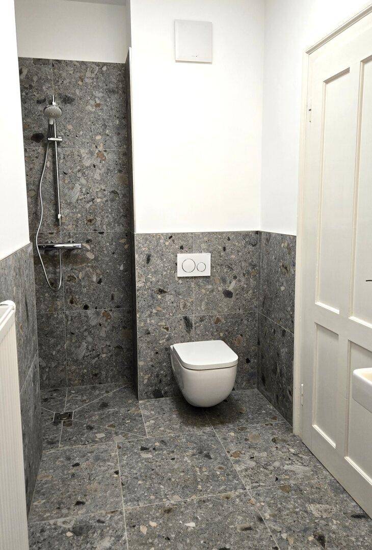 Modernes Badezimmer mit grauen Terrazzo-Fliesen, wandhängender Toilette und einer geräumigen begehbaren Dusche.
