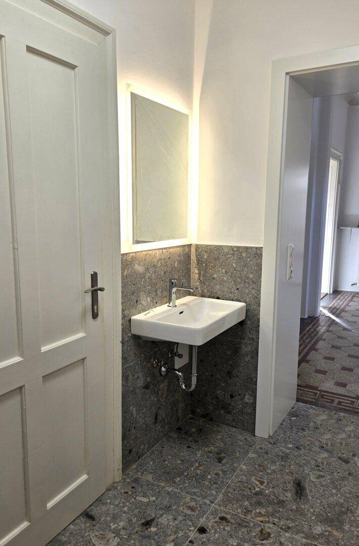 Badezimmer mit modernem Waschbecken, beleuchtetem Spiegel und stilvollen grauen Terrazzo-Fliesen an den Wänden.