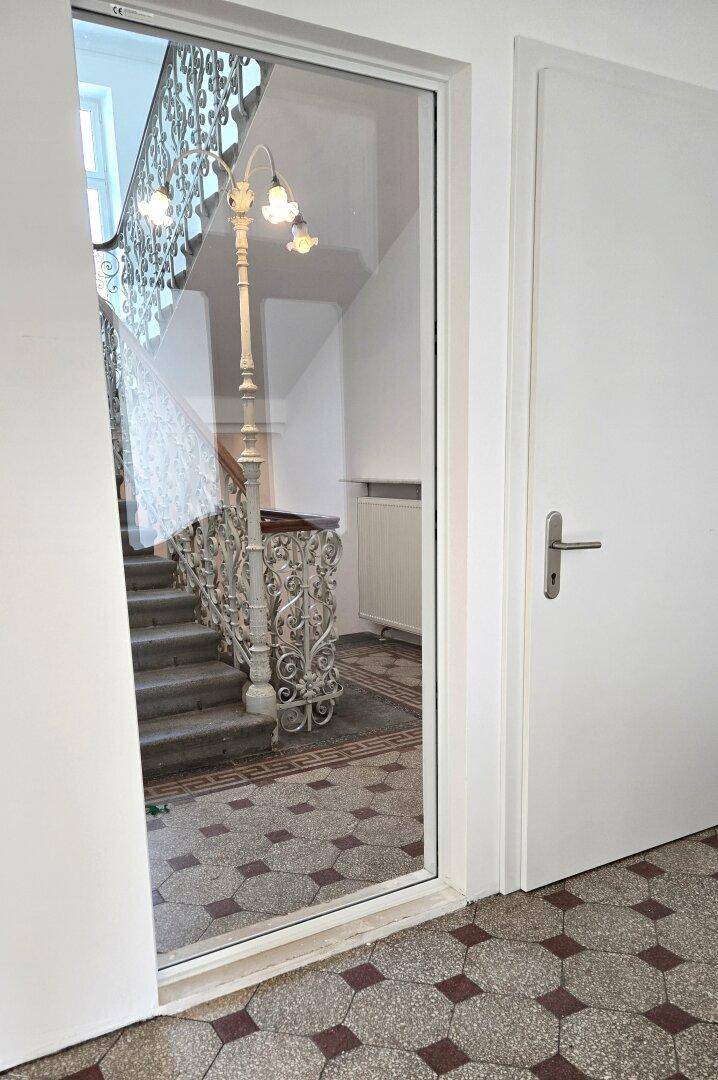 Blick durch eine Glastür auf ein historisches Treppenhaus mit kunstvollem Geländer und gemustertem Fliesenboden.