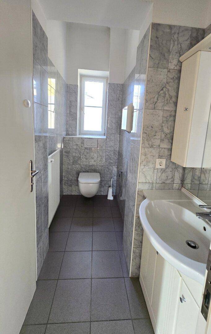 Gefliestes Badezimmer mit Fenster, Toilette und einem Waschbecken mit Unterschrank in hellem Grau.