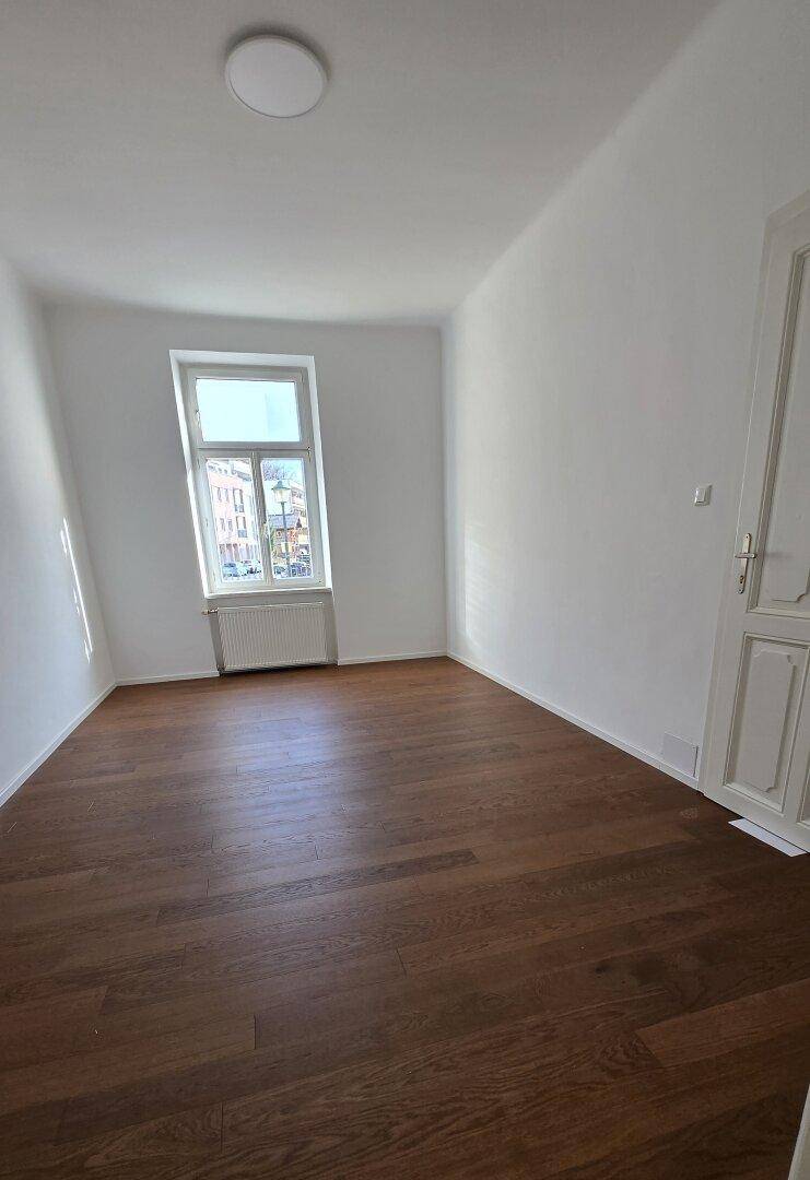 Helles Schlafzimmer mit dunklem Holzplankenboden, weißen Wänden und einem hohen Fenster mit Blick nach draußen.