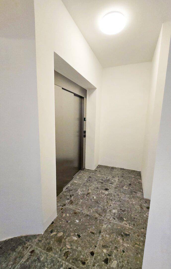 Moderner Flur mit grauen Terrazzo-Fliesen und einer eleganten Aufzugstür, die Zugang zu den Etagen bietet.