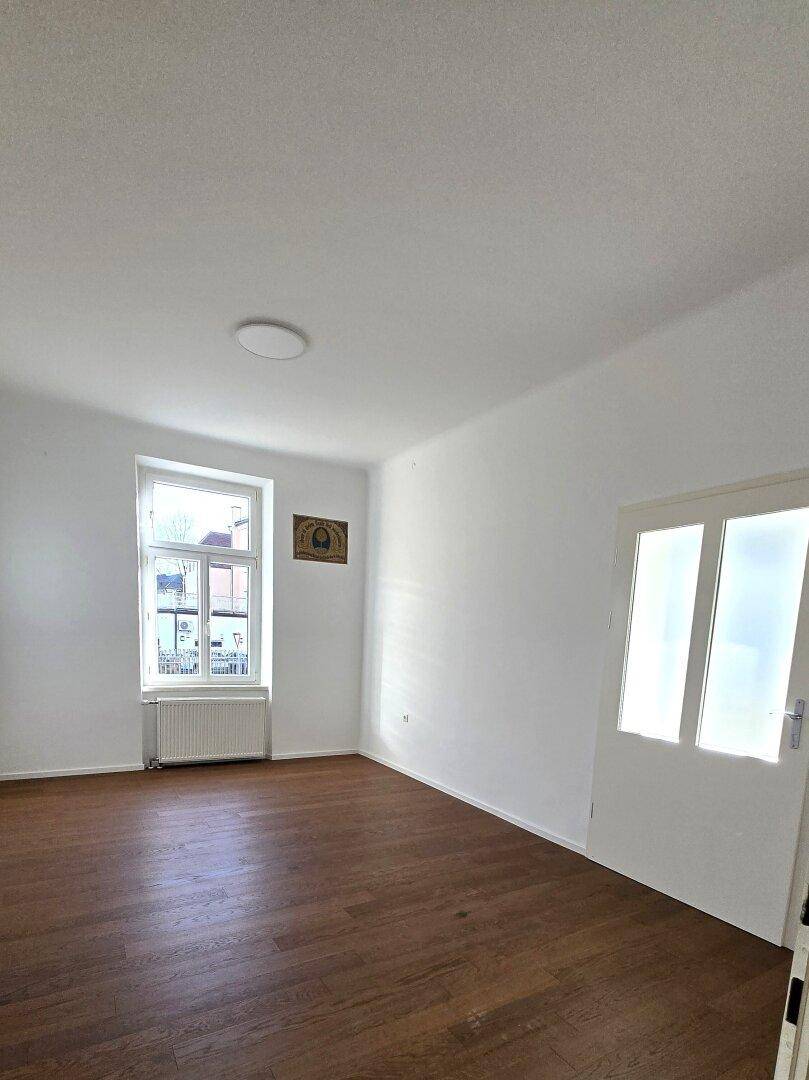 Helles, leeres Zimmer mit dunklem Holzplankenboden, weißen Wänden und einem hohen Fenster mit Blick nach draußen.