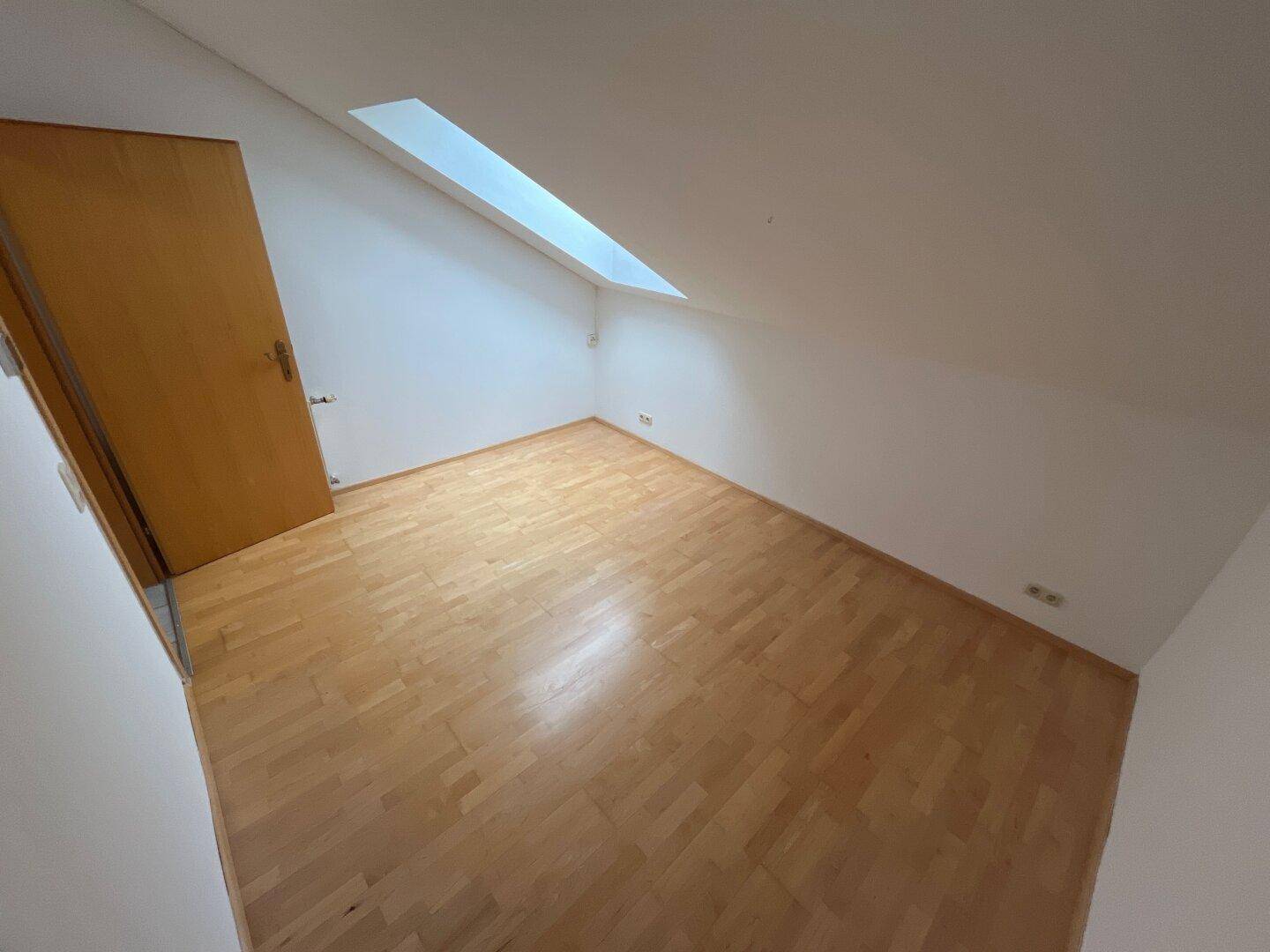 Schlichtes Zimmer mit Holzboden, Dachschräge und Dachfenster für natürliches Licht.