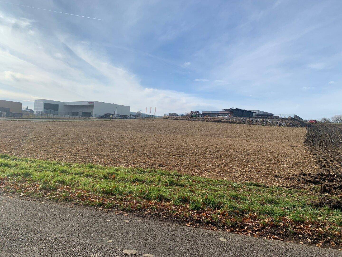 Weitläufiges Feld mit Ackerland und modernen Industriegebäuden am Horizont unter einem leicht bewölkten Himmel.