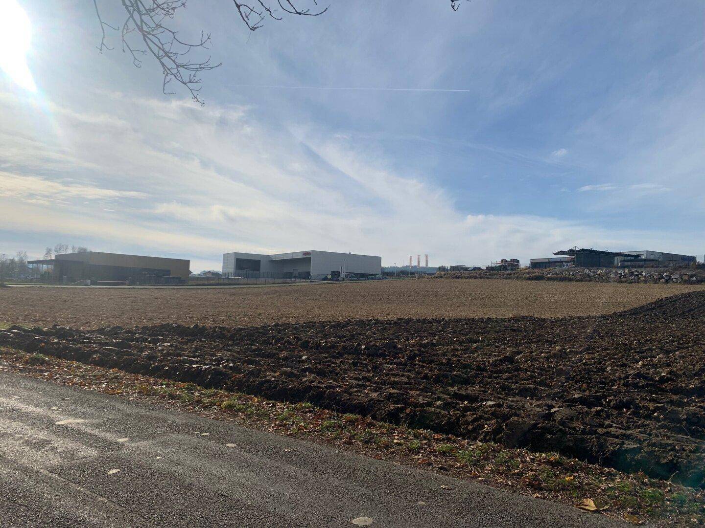 Weites Ackerland mit modernen Industriegebäuden in der Ferne unter einem blauen Himmel mit einigen Wolken.