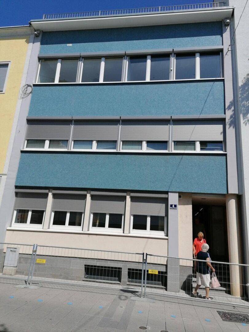 Mehrstöckiges Gebäude mit blauer Fassade, vielen Fenstern und Rollläden an einem sonnigen Tag.