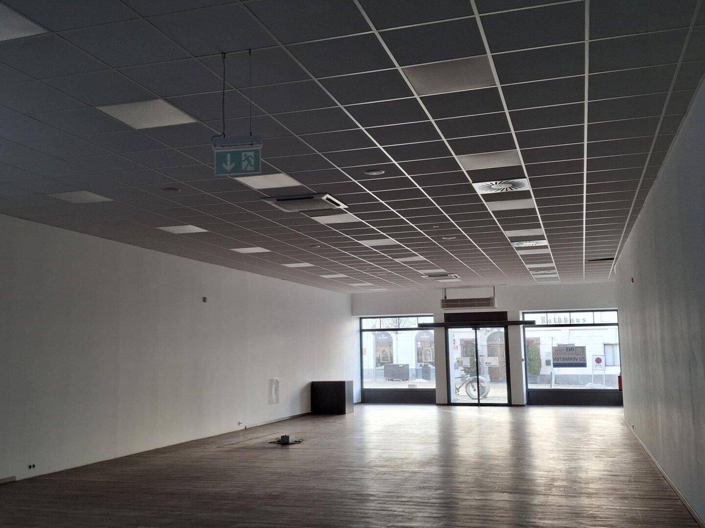 Großer, leerer Einzelhandelsraum mit Holzboden, abgehängter Decke und breiter Fensterfront für viel Tageslicht.