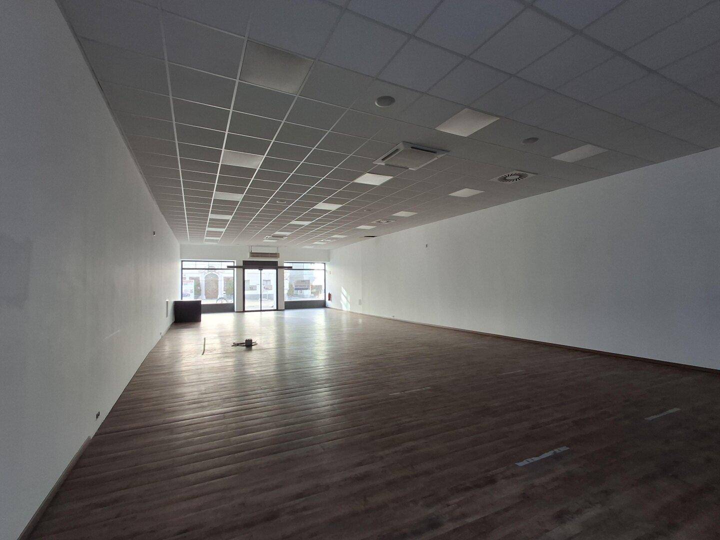 Weitläufiger, leerer Einzelhandelsraum mit Holzboden, weißen Wänden und viel Tageslicht durch die Fensterfront.