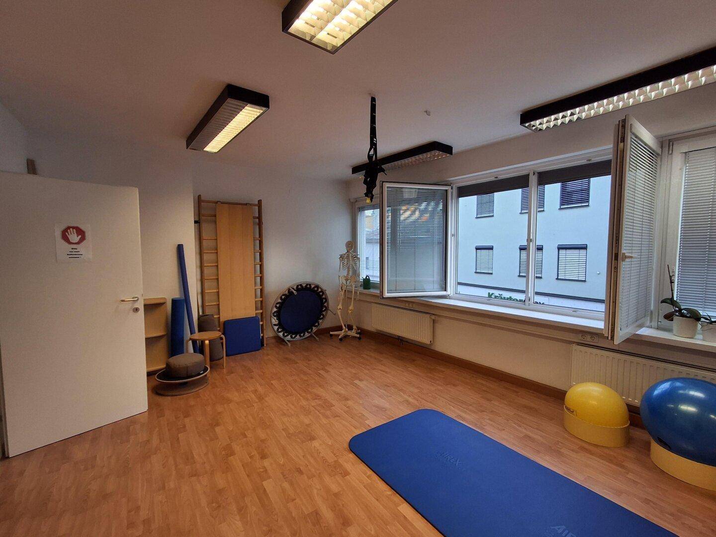 Heller Therapieraum mit Holzboden, Sprossenwand und medizinischen Modellen für Behandlungen und Training.