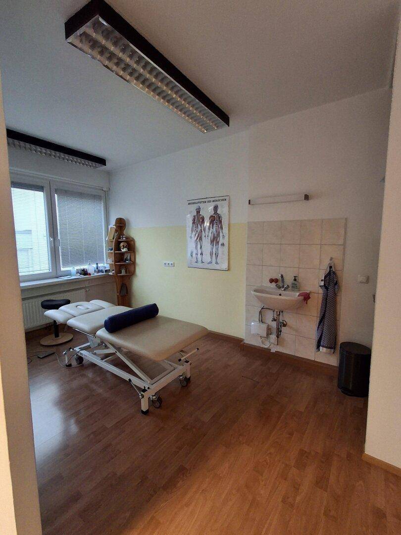 Behandlungsraum mit Therapieliege, Waschbecken und medizinischen Postern an der Wand für Patienten.