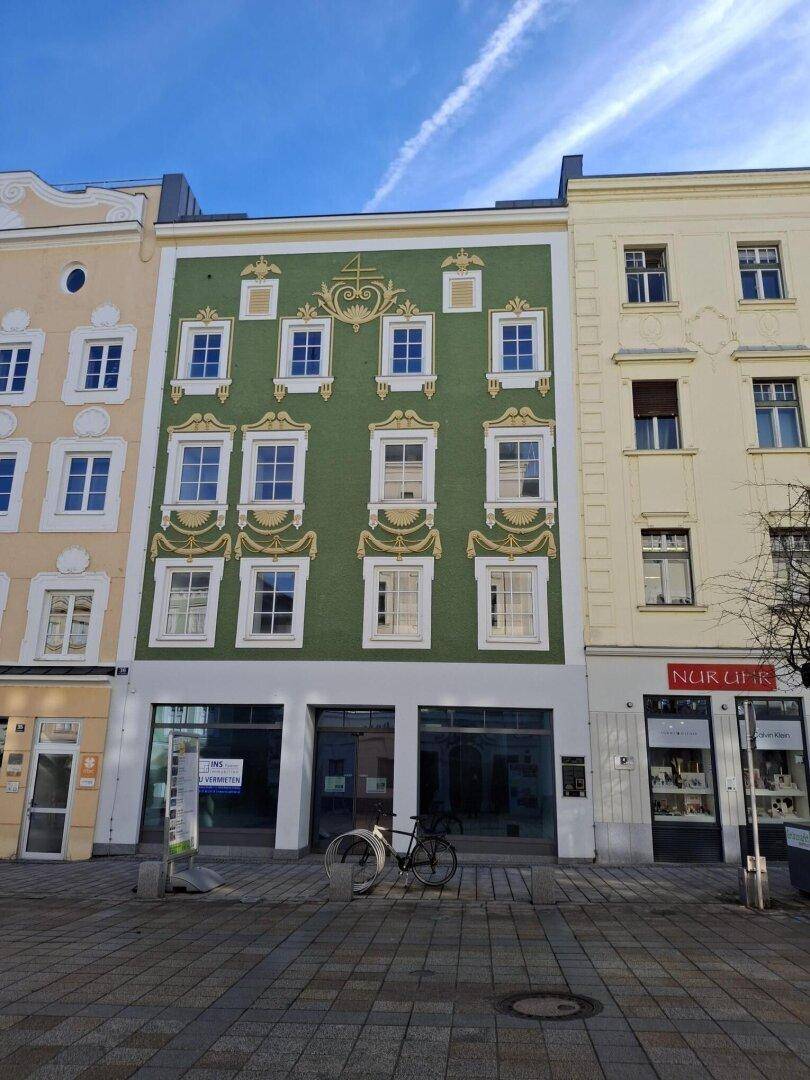 Historische Fassade eines Gebäudes mit grüner Farbe, weißen Fenstern und dekorativen Elementen. Im Erdgeschoss befinden sich Ladenflächen.