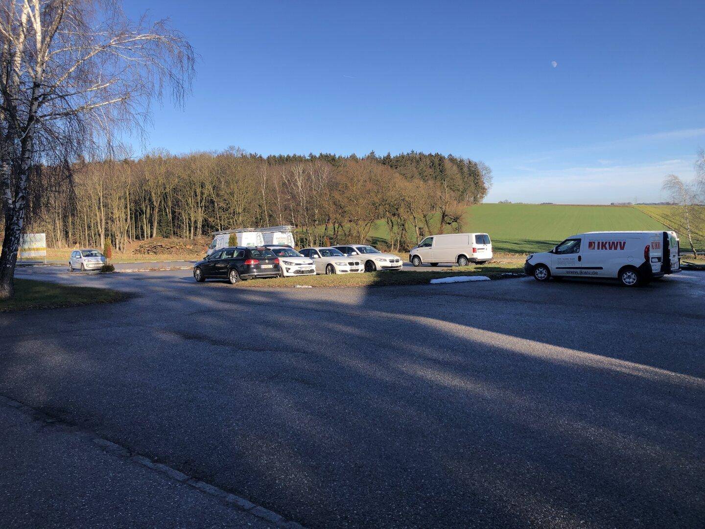 Parkplatz vor einem Waldgebiet mit mehreren Autos und Lieferwagen unter blauem Himmel.
