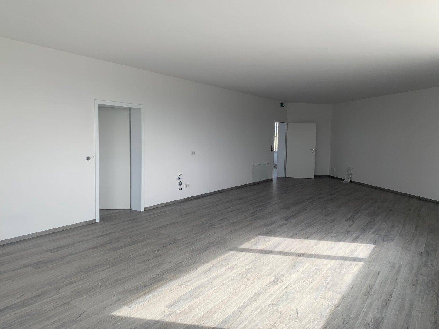 Heller Wohnbereich mit grauem Laminatboden und vorbereiteten Anschlüssen an der Wand.